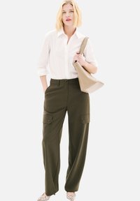Pantalons cargo vert olive avec poches latérales, associés à une chemise blanche à boutons. Le modèle tient un sac beige clair et porte des chaussures à motifs.