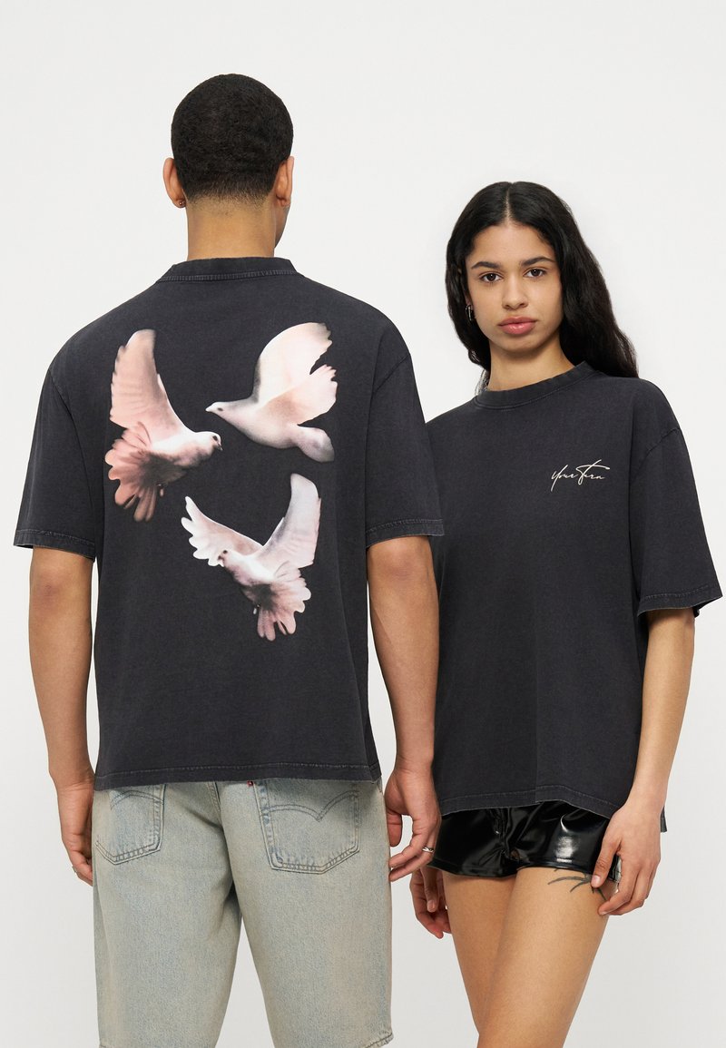 Deux personnes portant des t-shirts noirs oversize ; l'un affiche trois colombes blanches volant au dos, l'autre a une petite signature blanche à l'avant.