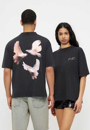 Deux personnes portant des t-shirts noirs oversize ; l'un affiche trois colombes blanches volant au dos, l'autre a une petite signature blanche à l'avant.
