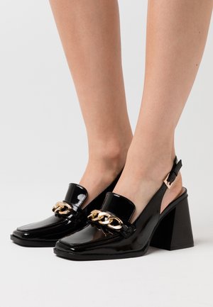 Sorte lakerede slingback-sko med en chunky hæl og guld kædeaccent på vampsen. Har en rund tå og skinnende overflade.