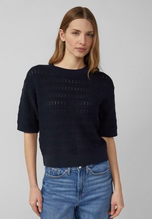 Kobieta ubrana w granatowy, krótki sweter z dzianiny z okrągłym dekoltem oraz jasnoniebieskie jeansy z wysokim stanem, stojąca na jednolitym szarym tle.