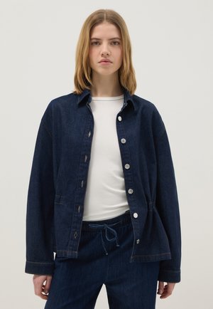 Junge Frau trägt eine dunkelblaue Jeansjacke über einem weißen Hemd und passende Jeanshosen, steht vor einem schlichten, hellen Hintergrund.