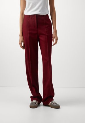 Nainen, jolla on ylipolvinen, suoralahkeinen viininpunaiset housut yhdistettynä harmaisiin ja pinkkeihin tennareihin, seisoo yksiväristä valkoista taustaa vasten.