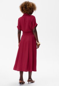 Dieprode jurk van zachte stof, met een kraagrand, korte oprolbare mouwen en een getailleerde taille met plooien die overgaan in een vloeiende rok.