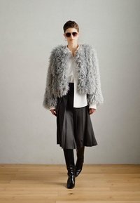 Kurtka z sztucznego futra w jasnoszarym kolorze, dopasowana biała koszula, plisowana czarna spódnica oraz czarne kozaki za kolano, zaprezentowane w minimalistycznym otoczeniu.