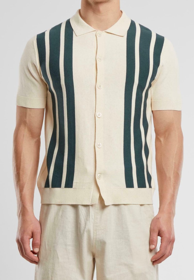 Homme portant une chemise crème à manches courtes avec des rayures verticales foncées et un pantalon crème assorti sur un fond uni.