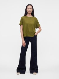 Femme debout, la main dans la poche, portant un haut olive à manches courtes, un pantalon large foncé et des chaussures pointues de couleur claire.