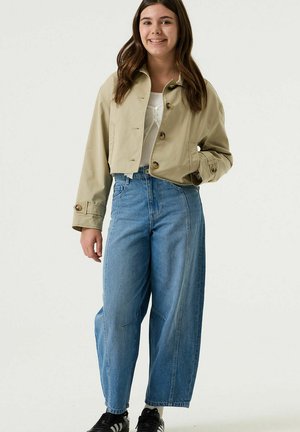 Junge Frau lächelt, trägt eine kurze beige Jacke, weißes Oberteil, lockere hoch taillierte blaue Jeans und schwarze Turnschuhe, steht vor einem schlichten Hintergrund.