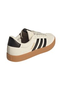 Vita mockasinsneaker med svart hälla, svart tredelad design, gummisula och märkesetikett på sidan med Adidas-logotyp.
