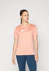Vaate t-paita, lyhythihainen ja pyöreäkauluksinen Nike, vaaleanpunaisesta kankaasta, jossa on suuri valkoinen Nike-logo rinnan kohdalla. Sileä tekstuuri, minimalistinen muotoilu.