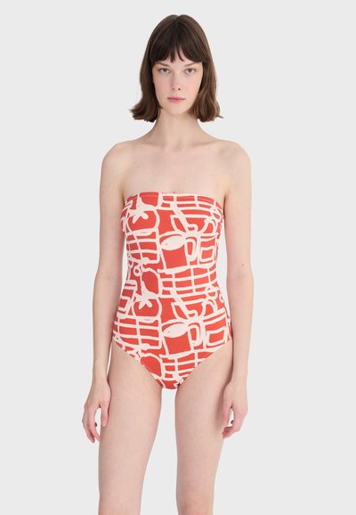 Femme aux cheveux bruns courts portant un maillot de bain orange sans bretelles avec un motif abstrait de fruits blancs et une grille, debout devant un fond clair uni.