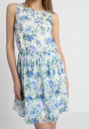 Bílých bezrukávových šatů po kolena s modro-zeleným květinovým vzorem, se staženým pasem, které nosí žena s dlouhými vlasy.