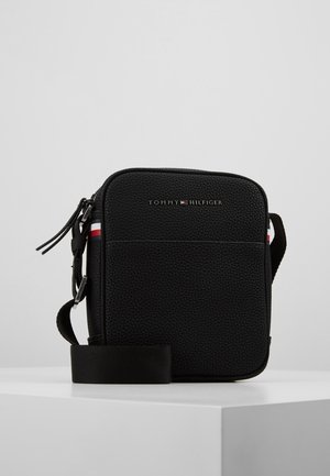Sac bandoulière noir texturé avec logo Tommy Hilfiger, accents en rayures rouge, blanc et bleu, et bandoulière réglable sur surface blanche.