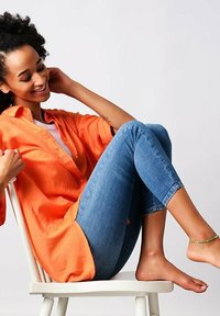 Løs orange skjorte over en hvid top, parret med tætsiddende blå jeans. Modellen sidder på en hvid stol, barfodet med et diskret ankelarmbånd.