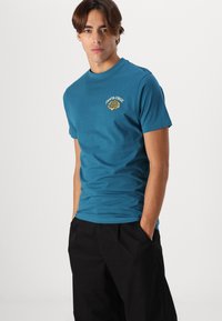 Turquoise cotton t-shirt featuring a yellow tiger graphic and text "SANTA CRUZ" on the left chest area. Short sleeves and crew neck style.