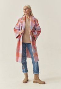 Villasegu seguga mantel, millel on suur roosa ja oranž ruuduline muster, koos beežiga kampsuniga, millel on karu kujundus, üleskeeratud teksade ja helepruunide pahkluuni saapadega.