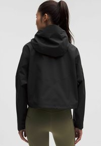 Kvinde med mørk hestehale iført en sort hættejakke med cropped design og olivengrønne tætsiddende leggings, stående med ryggen til på en ensfarvet baggrund.