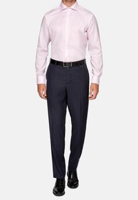 Man som bär en ljusrosa skjorta, mörkblå kostymbyxor med ett svart bälte och svarta polerade slipskor, stående mot en vit bakgrund.