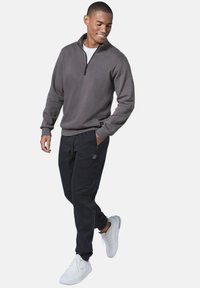 Grau zippe Pullover, schwarze Jogginghosen mit Reißverschlusstaschen, weiße Sneakers. Glatte Textur, lockere Passform, dezente Markenkennung auf der Kleidung.