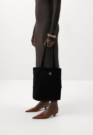 Personne portant une robe côtelée marron et un bracelet chaîne doré tenant un sac fourre-tout noir avec un petit motif blanc de soleil et lune. Talons pointus marron portés.