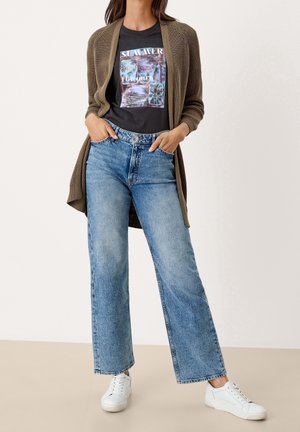 Cárdigan de punto abierto en color oliva sobre una camiseta gráfica negra con el texto "VERANO", combinado con jeans amplios de mezclilla azul desvanecido y zapatillas blancas.
