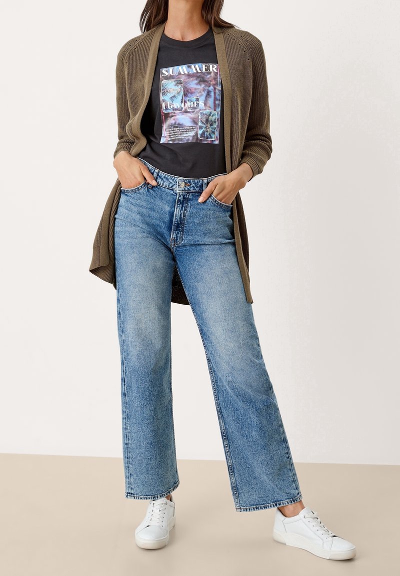 Olivfärgad öppet stickad cardigan över en svart t-shirt med grafiskt tryck som säger "SUMMER", ihop med vida, urblekta blå jeans och vita sneakers.