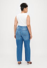 Blue high-waisted jeans with a straight cut, light fading, and two back pockets, paired with a white sleeveless top and heeled shoes.