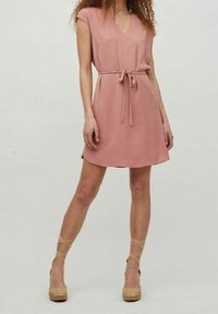 Robe rose sans manches avec un col en V et une ceinture à nouer à la taille, présentant une coupe ample et un tissu lisse. Associée à des espadrilles compensées.