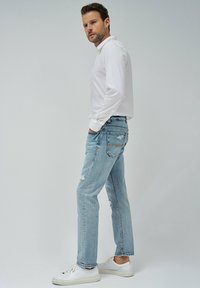 Jeans de mezclilla azul claro con un ajuste relajado, que presentan detalles desgastados. Combinados con una camisa blanca de manga larga y zapatillas blancas.
