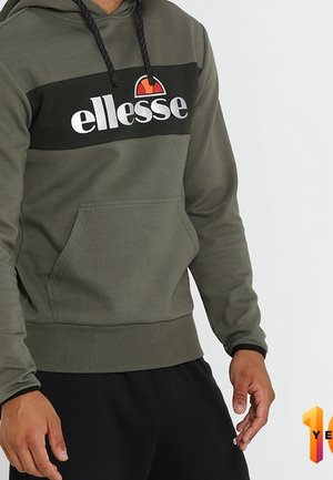 Mężczyzna w oliwkowo zielonej bluzie z kapturem marki ellesse z czarnymi sznureczkami i czarnych spodniach, stojący na tle jasnego, jednolitego tła.