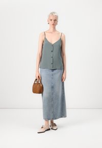 Top de tirantes verde claro con botones, combinado con una larga falda de mezclilla. Bolso de mano tejido en marrón y bailarinas beige con tiras decorativas.