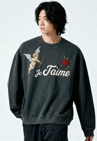 Jeune homme portant un sweat-shirt foncé avec une broderie représentant un cupidon, un cœur rouge et le texte "Je T'aime" sur la poitrine, regardant sur le côté.