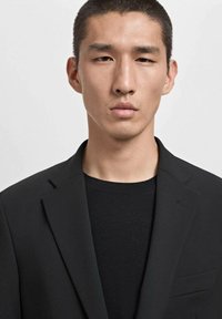 Blazer nero con revers a punta, chiusura a un bottone e tasca sul petto, indossato sopra un maglione a maglia nera con texture. Design pulito e sartoriale.