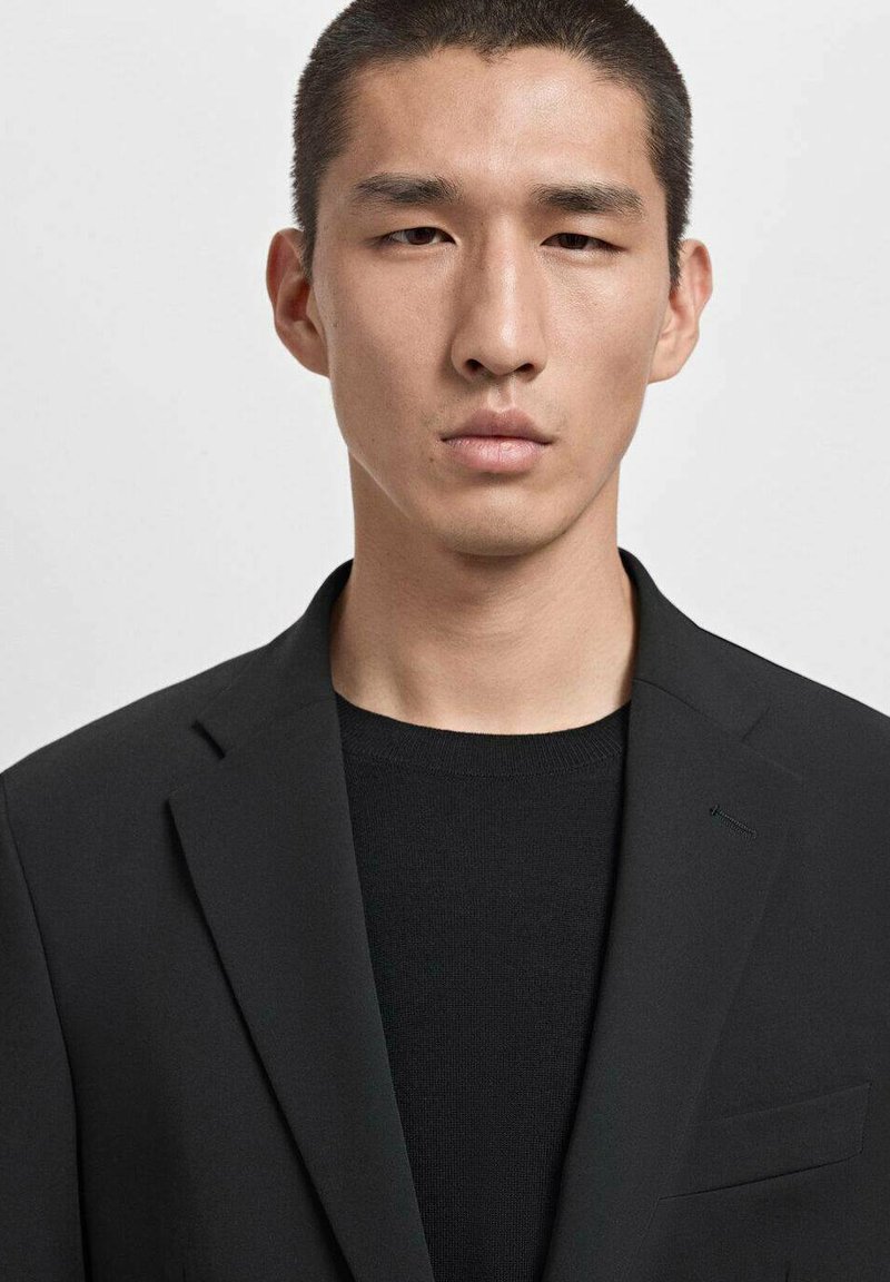 Blazer negro con solapas con muesca, cierre de un botón y un bolsillo en el pecho, usado sobre un suéter de punto texturizado negro. Diseño limpio y entallado.