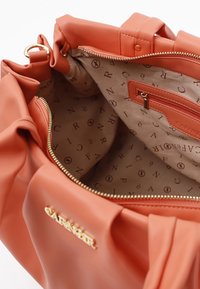 Perzikkleurige tote bag met een zachte, gladde textuur. Interieur heeft een beige voering met bedrukte logo's en een ritsvak voor opslag.