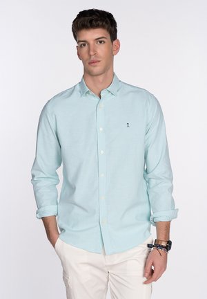 Camisa de manga larga color azul claro con un corte relajado, cuello abotonado y un logo bordado en el pecho. La tela parece texturizada y suave.