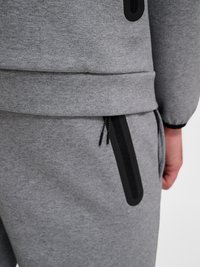 Nærbilde av en person som har på seg en grå genser og bukser med svarte glidelåsdetaljer på lommen og ermkanten.