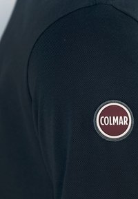 Tessuto nero testurizzato con una cucitura arrotondata sulla spalla e una toppa circolare maroon con la scritta "COLMAR" sul braccio.
