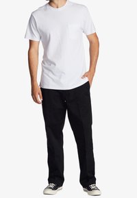 Biały bawełniany t-shirt z okrągłym dekoltem i krótkimi rękawami, z kieszonką po lewej stronie na piersi, zestawiony z czarnymi spodniami i czarnymi trampkami.