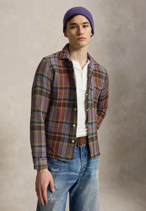 Jeune homme portant un bonnet tricoté violet, une chemise à carreaux multicolores par-dessus un henley blanc, et un jean bleu éclaboussé de peinture avec une ceinture marron.
