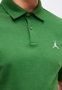 Polo vert avec deux boutons fermés et un petit logo en silhouette blanche d'un joueur de basket-ball sur la poitrine.
