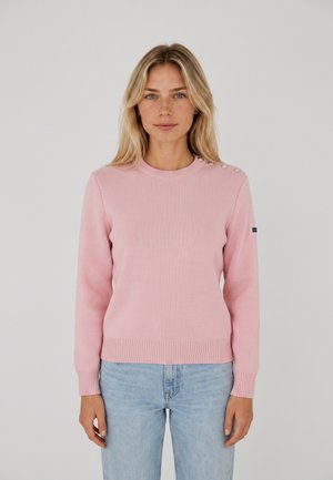 Donna con capelli biondi lisci che indossa un maglione rosa a maniche lunghe con bottoni sulla spalla e jeans azzurri chiari, in piedi contro uno sfondo semplice.