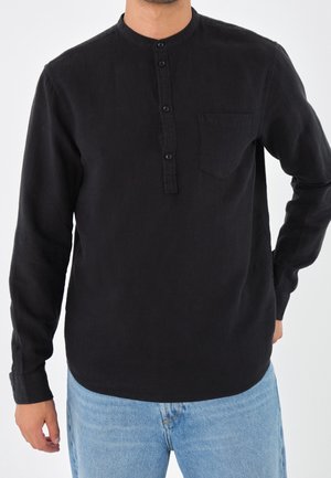 Mand iført en sort langærmet Henley-trøje med knapper og brystlomme, kombineret med lyseblå jeans, stående mod en hvid baggrund.