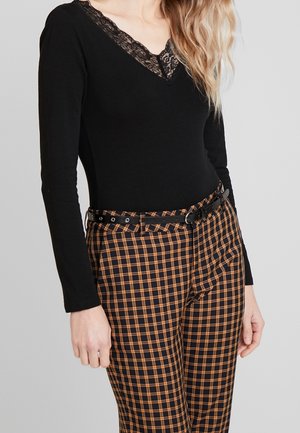 Femme portant un haut noir à manches longues avec un col en V en dentelle et un pantalon taille haute à carreaux orange et noir avec une ceinture noire.