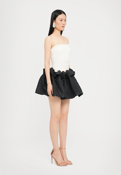 Top blanco sin tirantes con detalles fruncidos, combinado con una falda negra de satén voluminoso. La modelo lleva sandalias desnudas con tiras y grandes pendientes blancos.