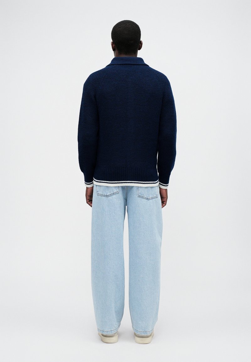 Navy gebreide trui met een relaxte pasvorm, voorzien van een geribbelde onderrand en manchetten. Gecombineerd met ruime lichtblauwe spijkerbroek en witte sneakers.