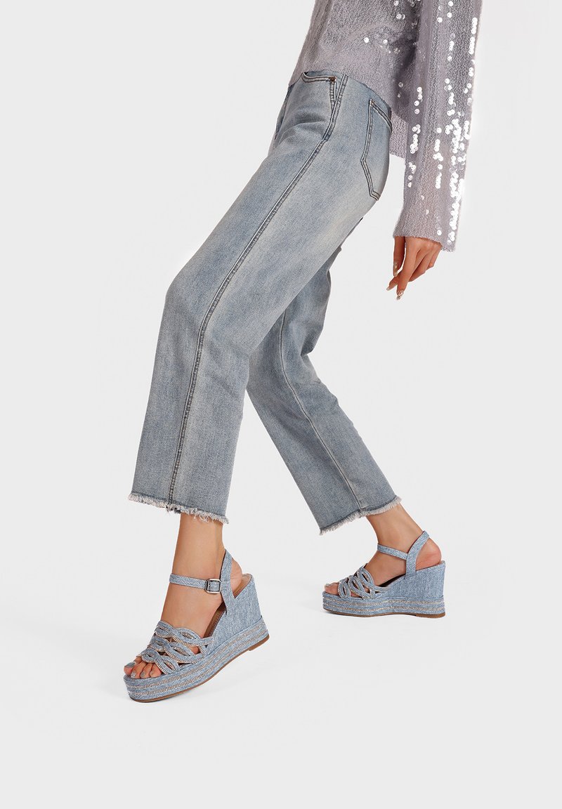 Sandali con zeppa in denim azzurro chiaro con design a tracolla intrecciata, fibbia regolabile alla caviglia e piattaforma testurizzata. Abbinati a jeans sfilacciati.