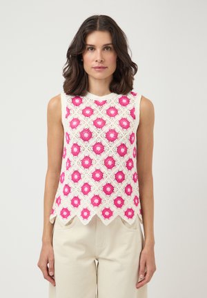 Kobieta ubrana w białą, bezrękawnikową bluzkę na szydełku z różowymi wzorami kwiatów i falowanym brzegiem, w połączeniu ze spodnmi w jasnobeżowym kolorze.