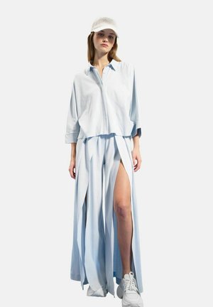 Femme portant une chemise ample bleu clair et une longue jupe fendue, des baskets blanches et une casquette blanche en filet, debout devant un fond uni.