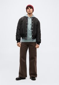 Νέος άνδρας που φοράει κόκκινο σκουφάκι, μαύρο μπουφάν τύπου bomber με κουμπιά, γκρι φούτερ με κουκούλα, καφέ φαρδιά παντελόνια και μαύρα παπούτσια, στέκεται μπροστά σε απλό φόντο.